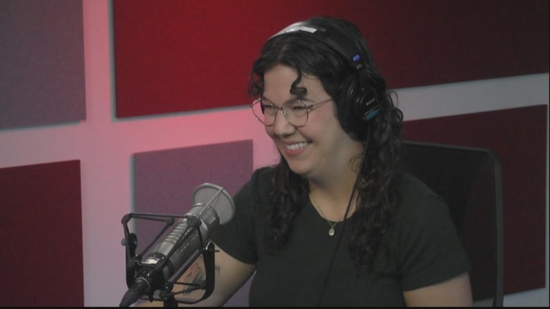 A smiling dark-haired woman, wearing a black shirt and glasses. He is sitting at the desk in front of the microphone.