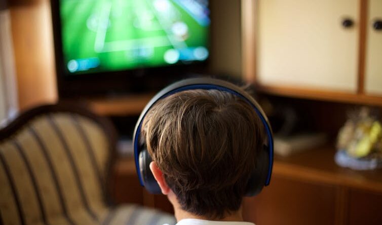 Back of the head of a boy wearing headphones and looking at a TV screen.