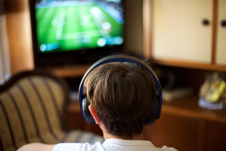 Back of the head of a boy wearing headphones and looking at a TV screen.